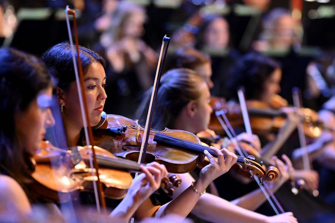 A group of female students, wearing smart attire, playing the violin in an orchestra performance.